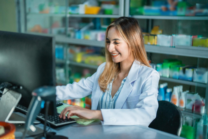 Pharmacist smiling while using the computer in a modern pharmacy, processing an online Juvederm order.