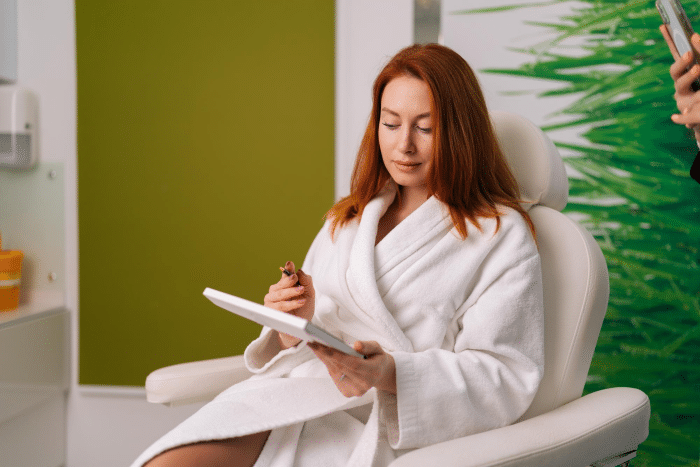 Red-haired woman in a spa robe filling out a consent form before a Dermalax filler treatment, representing a client's preparation step and the cost-related considerations involved
