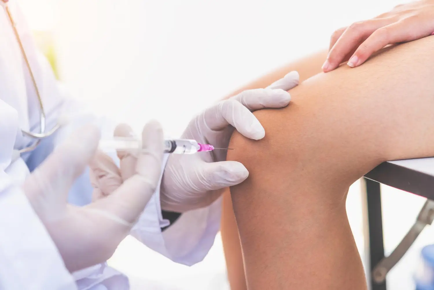 A medical professional in a white coat and gloves administers an injection to a patient's knee. 