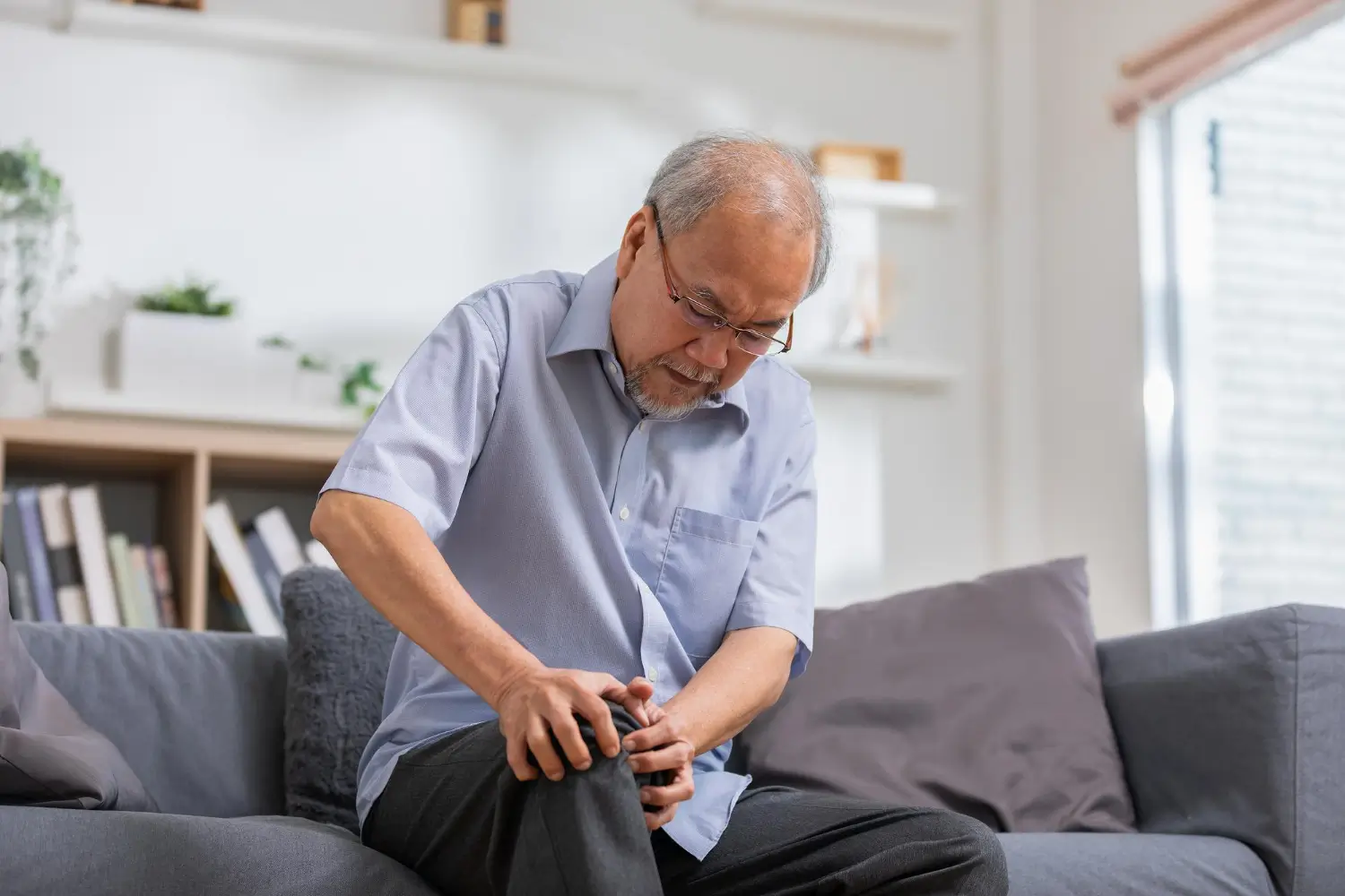A man sits on a gray sofa in a bright living room, massaging his knee with a slight expression of pain. 