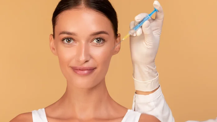 A woman with dark hair is smiling and looking at the camera. A gloved hand is holding a syringe near her face. The background is a warm peach color. She is wearing a white top.