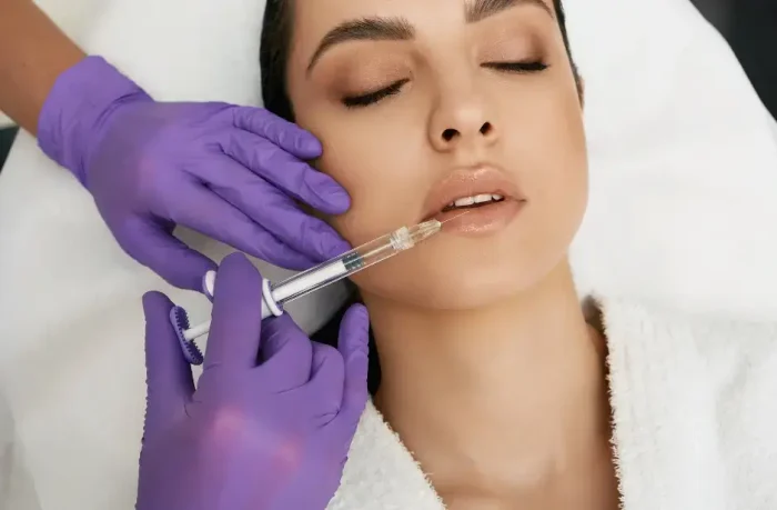 A person receiving a cosmetic injection in the lips while lying down, administered by someone wearing purple gloves. The individual appears relaxed, with eyes closed, wearing a white robe.