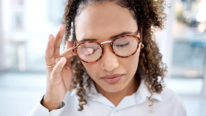 A person with curly hair and glasses appears to have their eyes closed, touching the side of their head gently with one hand. They are wearing a white shirt, and the background is softly blurred.