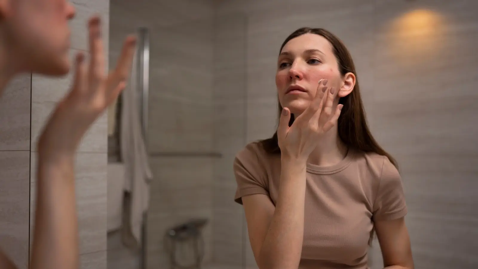 A woman in a beige shirt stands in a bathroom applying cream to her cheek. She looks at her reflection in the mirror. The bathroom has a towel hanging and beige tiled walls.