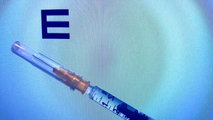 A close-up of a syringe with a needle and a capped tip, positioned diagonally against a blue background. Large, bold letter "E" partially visible in the top left corner.