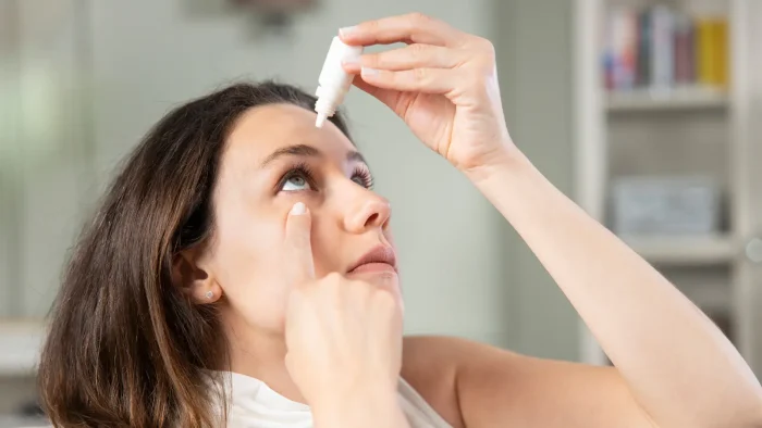 A woman with long brown hair is tilting her head back and applying eye drops to her left eye. She is indoors and wearing a white top, with a blurred background featuring shelves and decorative items.