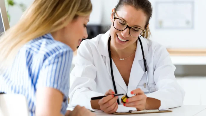 A doctor wearing glasses and a white lab coat discusses a prescription bottle with a patient. They are both seated at a desk, smiling and engaged in conversation. The doctor points to the bottle with a pen. Office supplies are in the background.