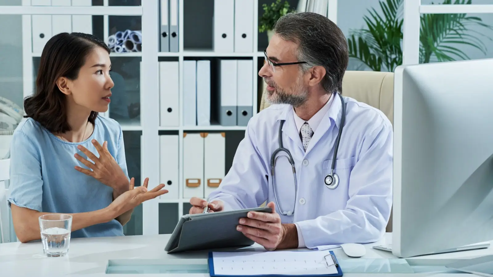 A doctor, seated at a desk with a stethoscope around his neck, holds a tablet while speaking to a woman sitting across from him. She gestures with her hands as they engage in conversation. Shelves with binders and plants are in the background.