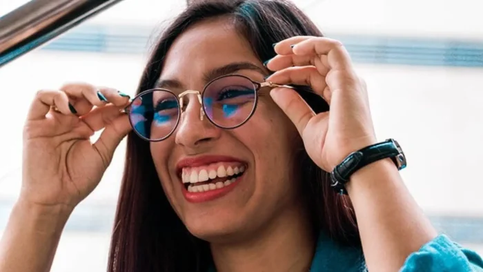 A smiling person with long hair adjusts their glasses. They are wearing a blue shirt and a black watch, with a blurred background.