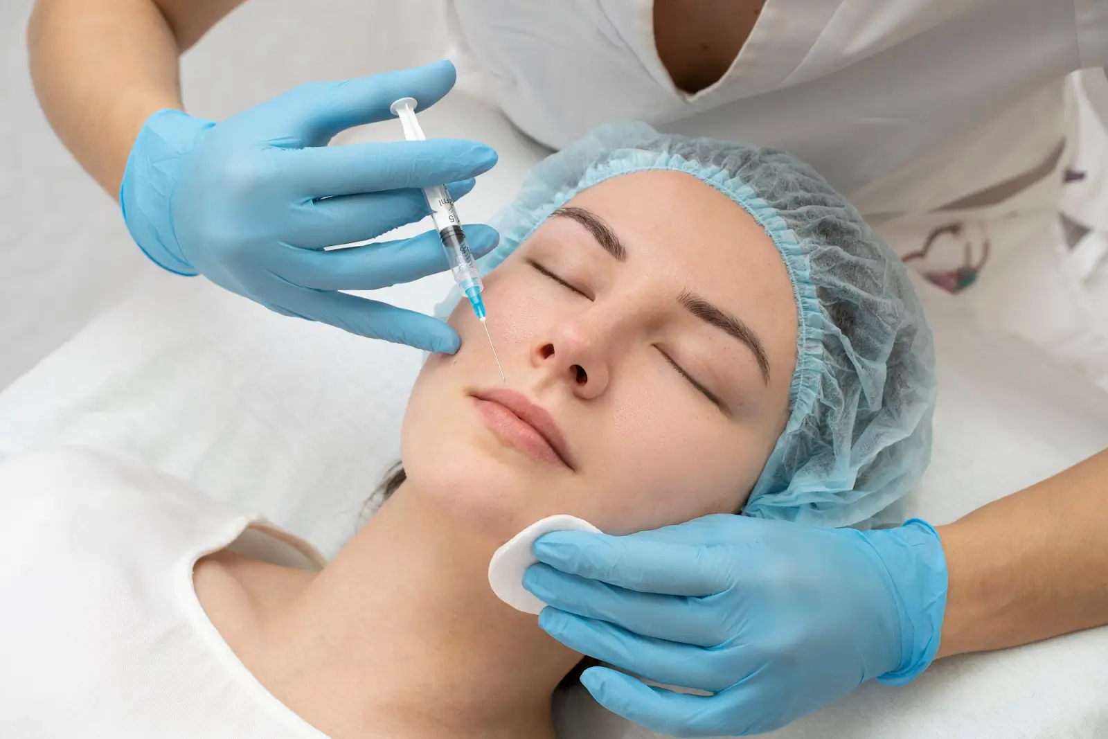 A person is receiving an injection in the face while lying down. They are wearing a hair cap, and a practitioner with blue gloves is holding a syringe and a cotton pad. The setting appears to be a clinical or spa environment.