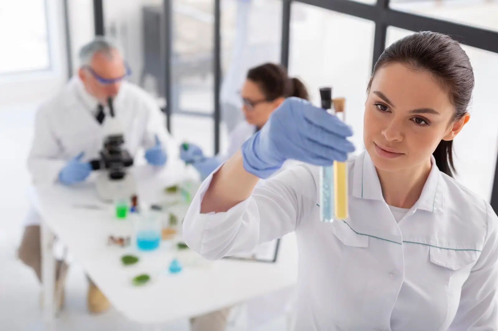 A woman in a lab coat and blue gloves examines a test tube filled with yellow liquid. In the background, two colleagues, also in lab coats, work at a table with microscopes and various lab equipment. The setting is a bright laboratory.