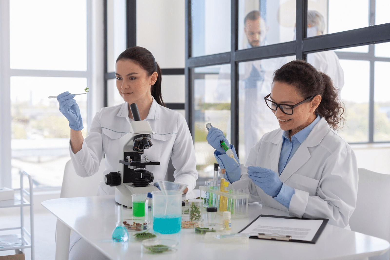 Two scientists in lab coats and gloves are working in a laboratory. One is using a pipette, and the other is examining a sample. A microscope, test tubes, and beakers with colored liquids are on the table. A third person is in the background.