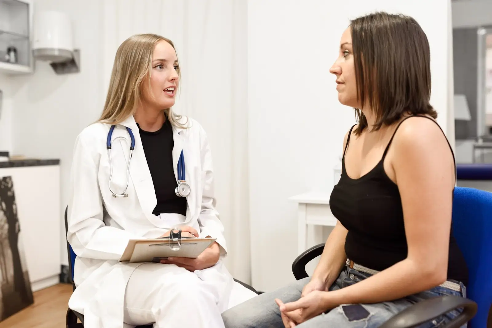 A doctor in a white coat, holding a clipboard, is sitting and talking to a patient in a medical office. The patient, wearing a black tank top, is attentively listening. Medical equipment and office furniture are visible in the background.