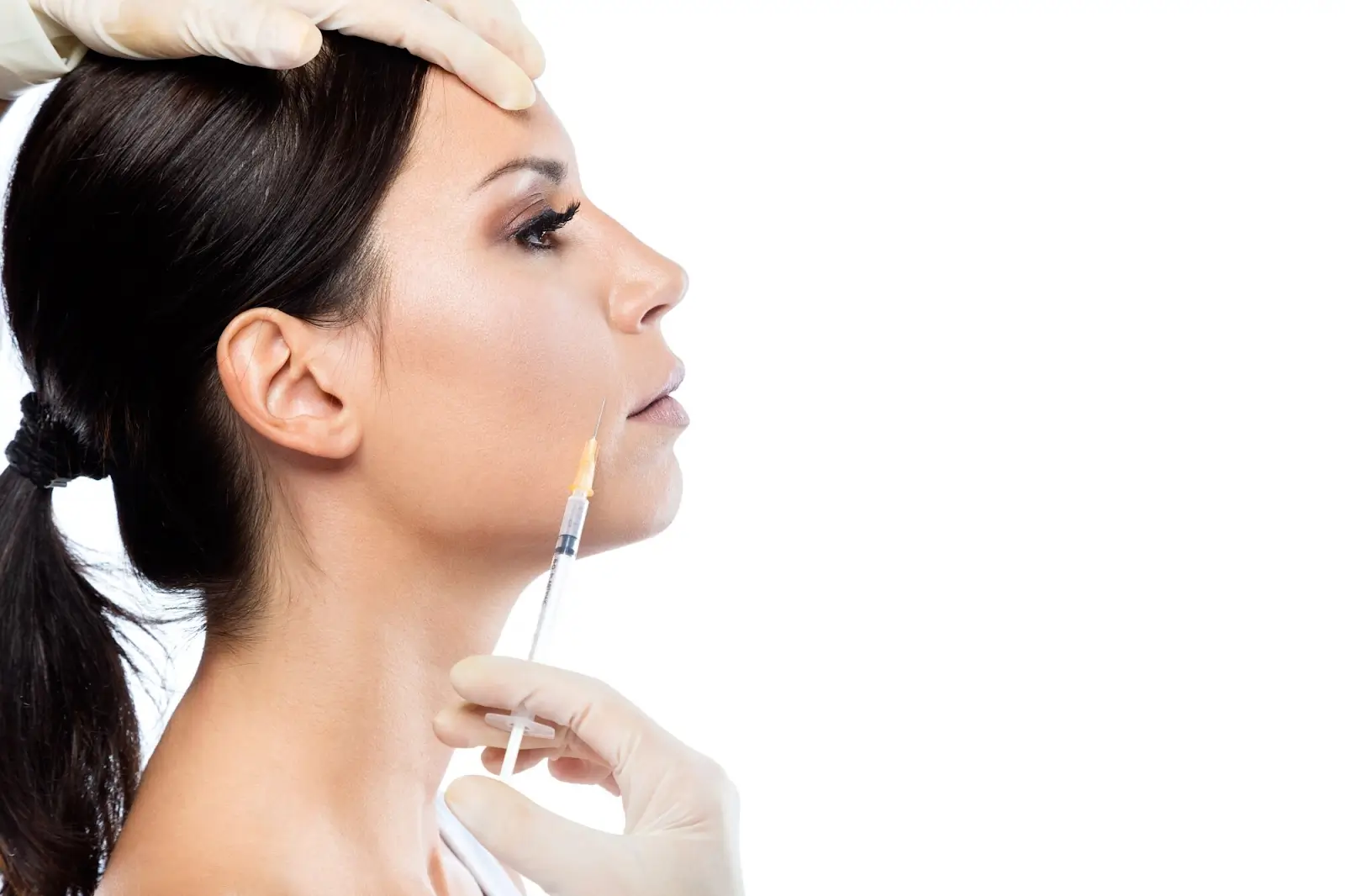 A woman with dark hair receives a cosmetic injection in her cheek from a gloved hand. Another gloved hand gently holds her head. The background is white, emphasizing the medical setting.