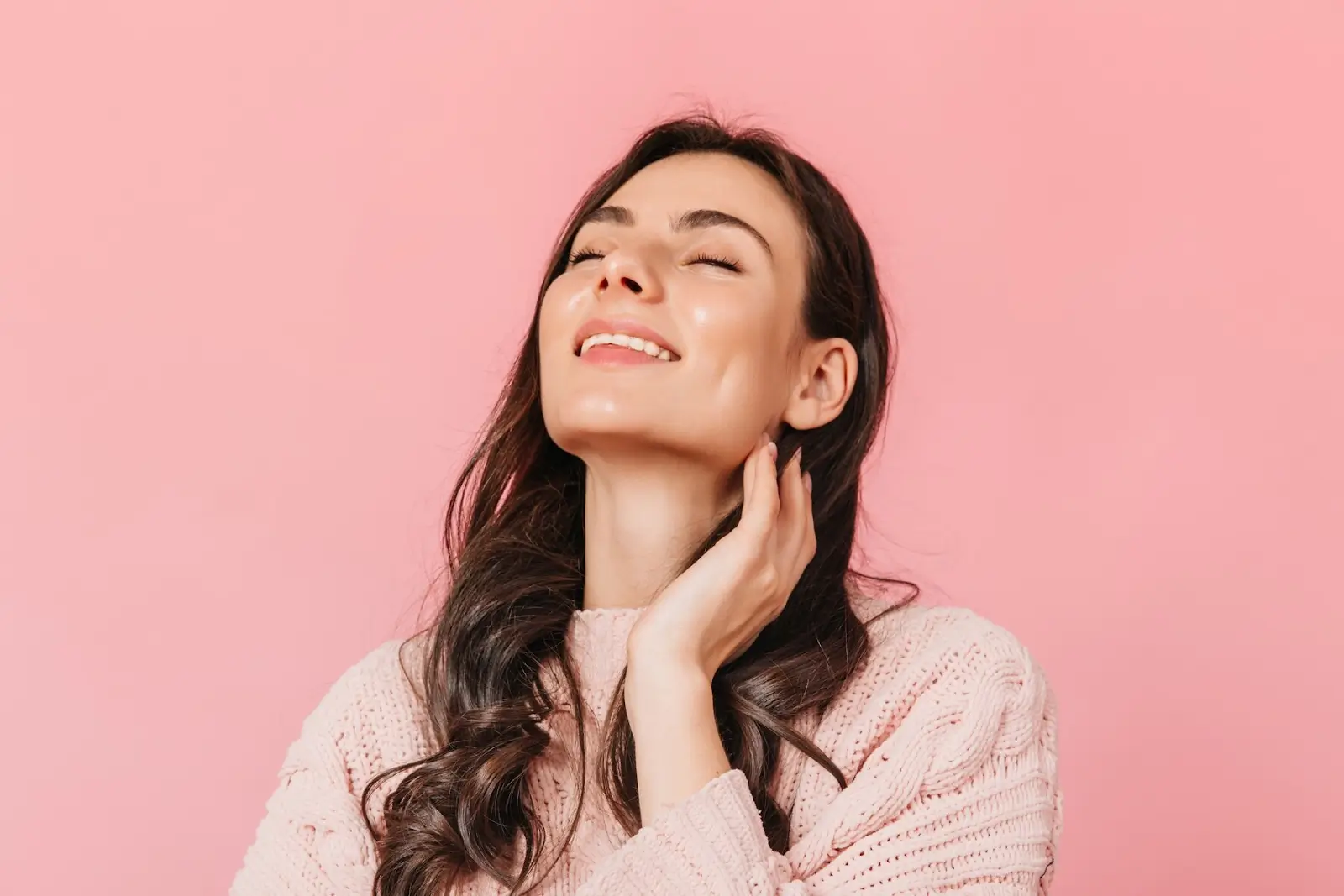 A woman with long, dark hair smiles with her eyes closed, tilting her head back and touching her neck. She is wearing a light pink sweater against a matching pink background.