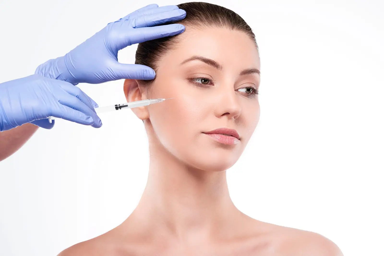 A person with bare shoulders receives a cosmetic injection in the cheek. A gloved hand holds the syringe against the skin, while another gloved hand gently touches the person's head. The background is plain white.