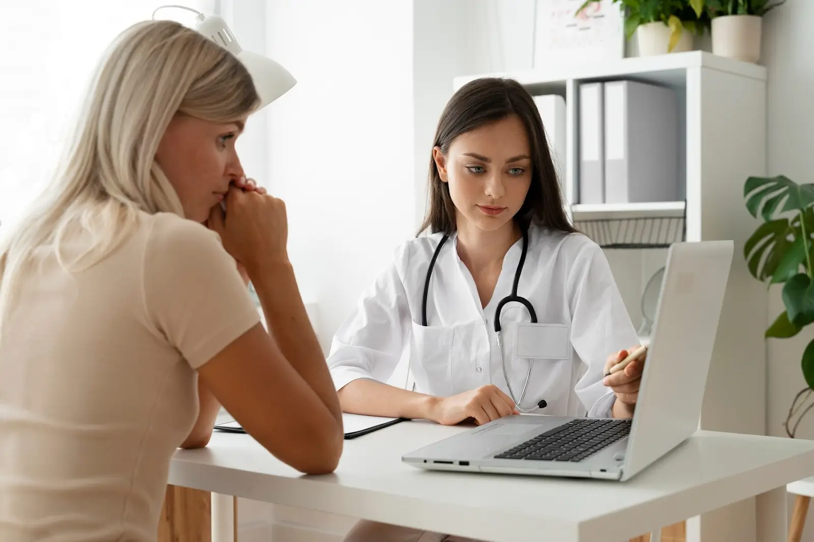 A doctor with a stethoscope discusses information on a laptop with a patient in a medical office. The patient listens attentively. The room is well-lit with plants and shelves in the background.