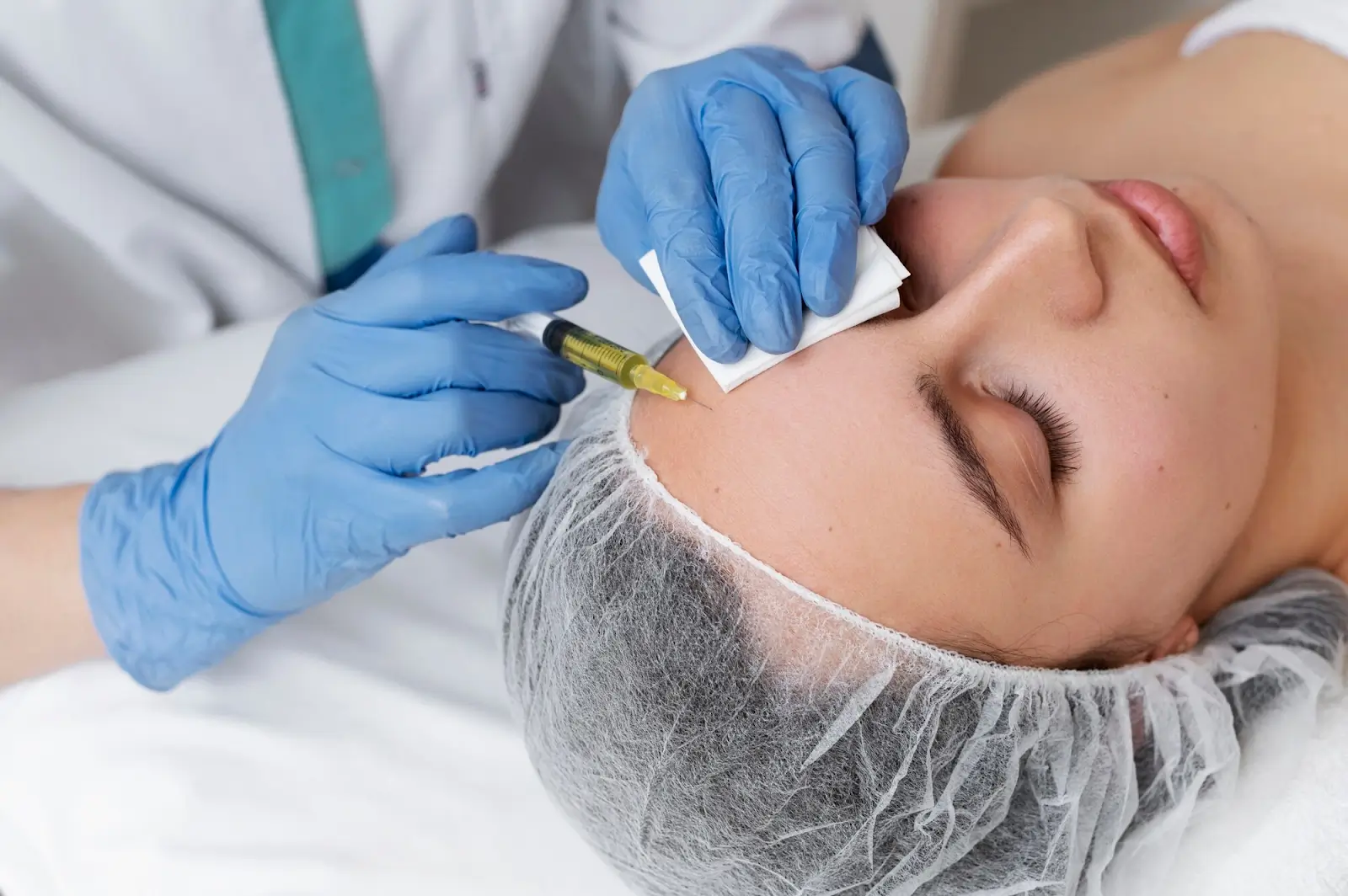 A person receiving an injection in the forehead by a medical professional wearing blue gloves. The recipient is lying down with eyes closed, covered with a hair net, while a white cloth is held on their forehead.
