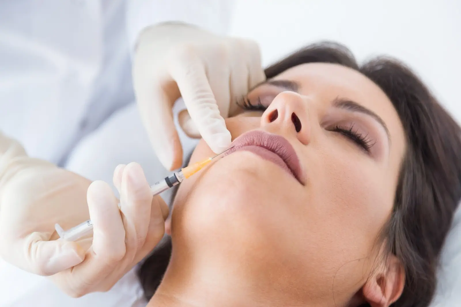 A person receiving a cosmetic injection in their lips. They are lying down with eyes closed. A gloved hand is holding the syringe, gently administering the procedure. The setting appears to be a clinical environment.