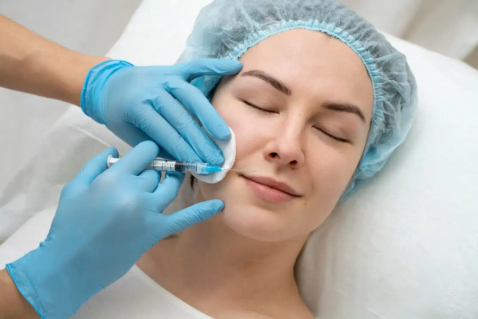A person lying down with eyes closed receives a facial injection from someone wearing blue gloves. The individual has a calm expression and is wearing a blue hair cover. The setting appears to be a medical or cosmetic treatment environment.
