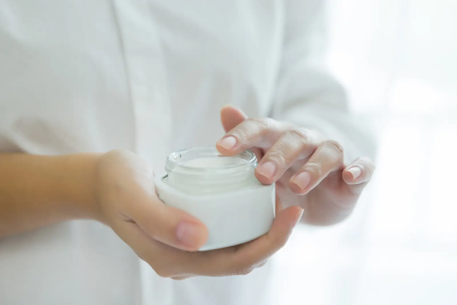 Close-up of a person in a white shirt holding an open white jar of cream in one hand, with the other hand's fingers ready to scoop from it. The background is softly blurred, creating a serene and clean atmosphere.