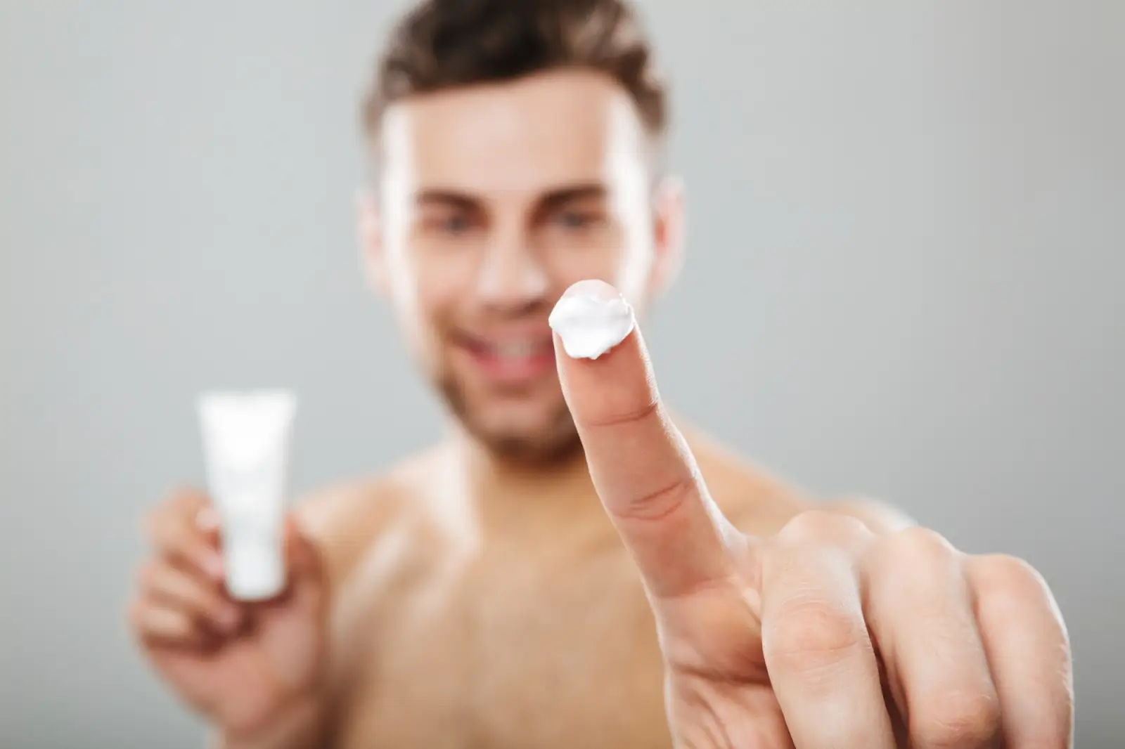 A man with short hair is smiling and holding a small white tube in one hand. His other hand is in focus, pointing at the camera with white cream on his fingertip. The background is a plain, light gray.