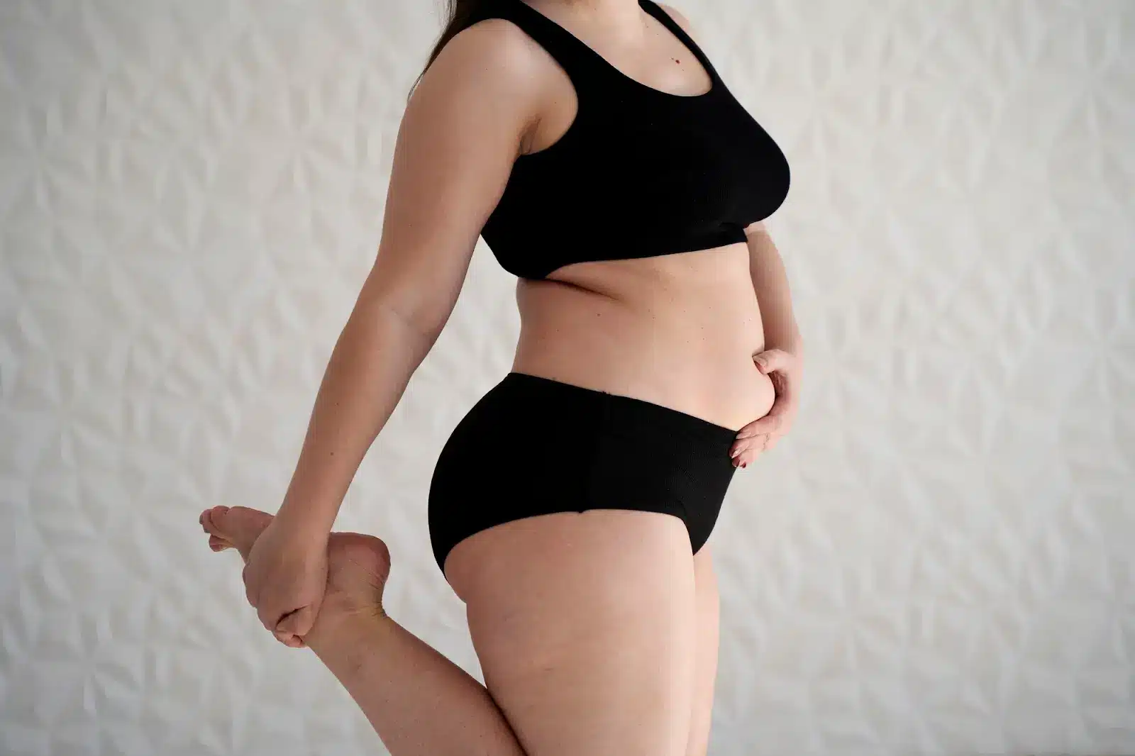 A woman in black underwear and a black sports bra stands on one leg, holding her other foot behind her in a stretch, against a light patterned background.