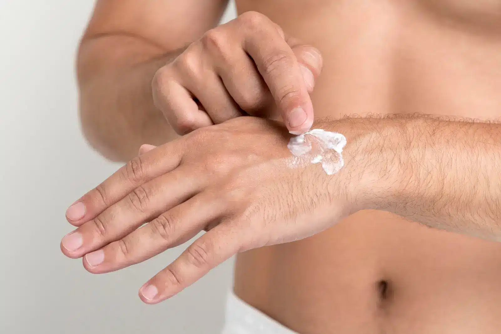 A person applying a small amount of cream onto the back of their left hand using their right fingers. The background is plain, highlighting the focus on skincare. The person is shirtless, emphasizing the attention on the hand and cream application.