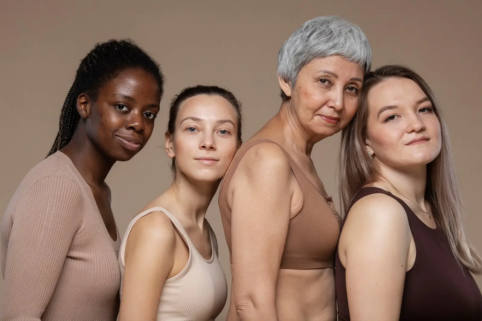 A group of four women stands closely together against a neutral background. They wear neutral-toned tops and display diverse ages, skin tones, and hair colors. They are smiling gently and looking at the camera.
