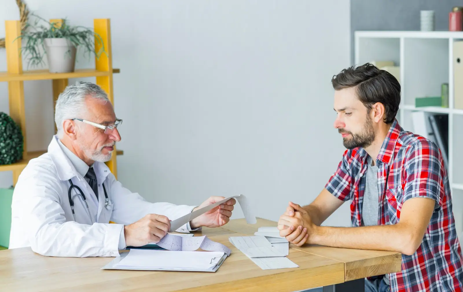 A doctor in a white coat sits at a desk reviewing papers with a young man in a red plaid shirt. They appear to be discussing medical information. The room is bright, with shelves and plants in the background.