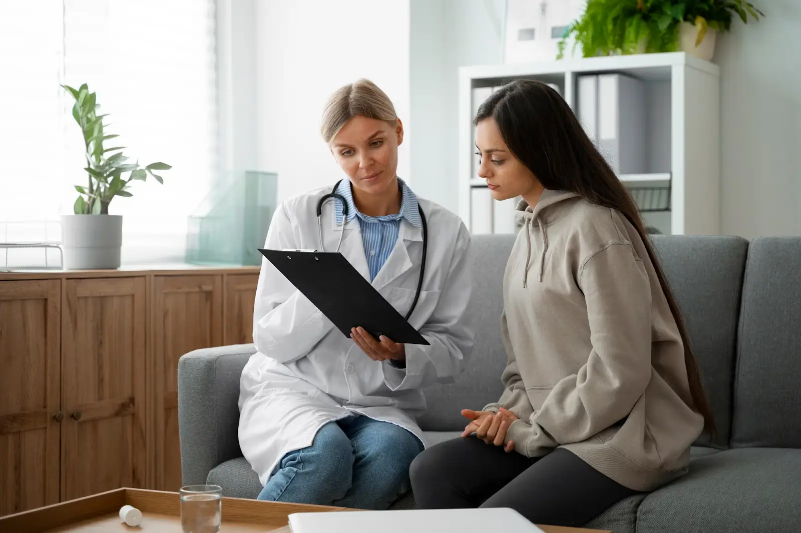 A female doctor in a white coat shows a clipboard to a young woman in a hoodie as they sit together on a sofa in a brightly lit office. The two appear to be having a serious conversation.