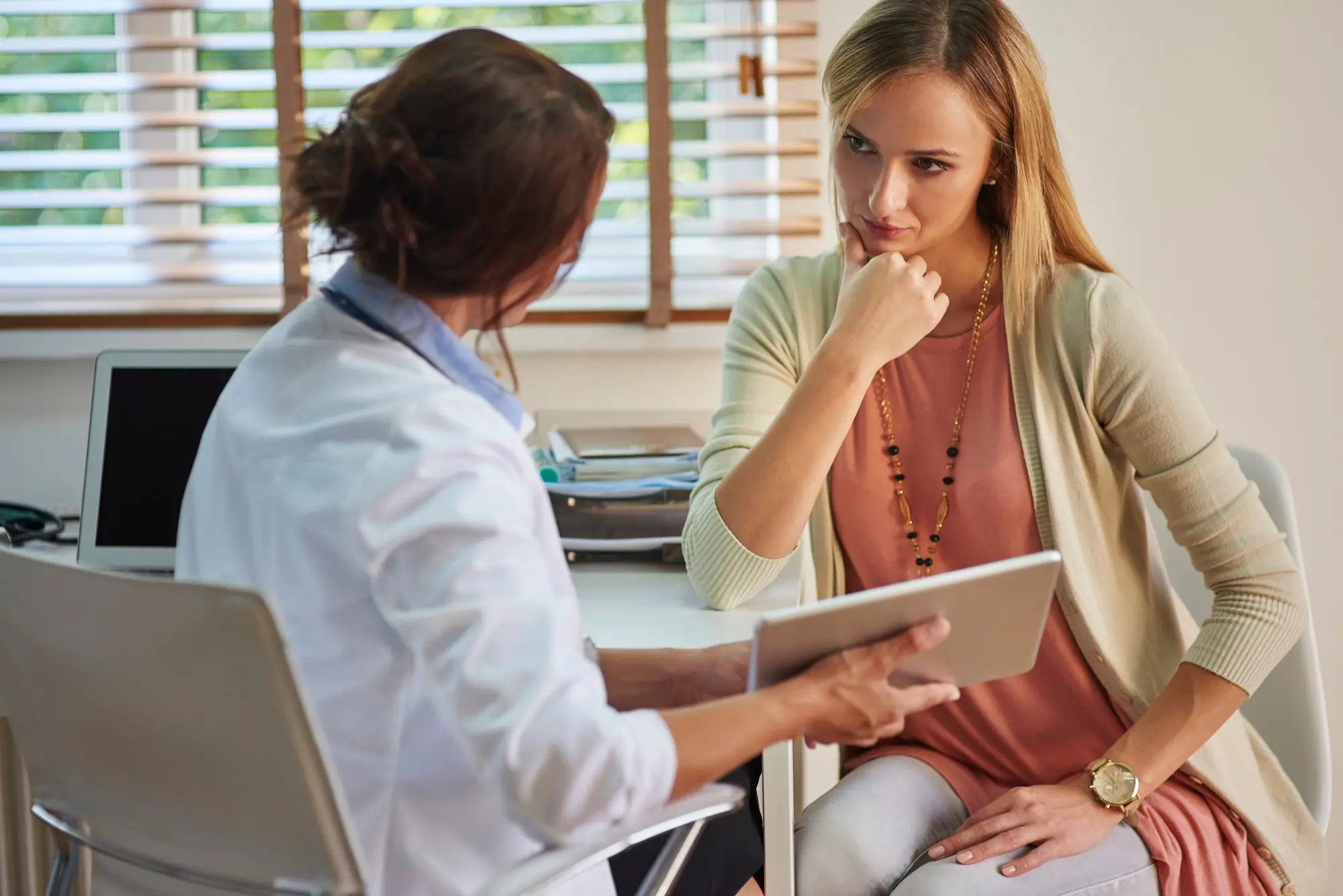 A doctor showing a patient information on a digital tablet. The patient listens attentively, resting her chin on her hand. They are seated in an office with a window in the background, partially covered by blinds.