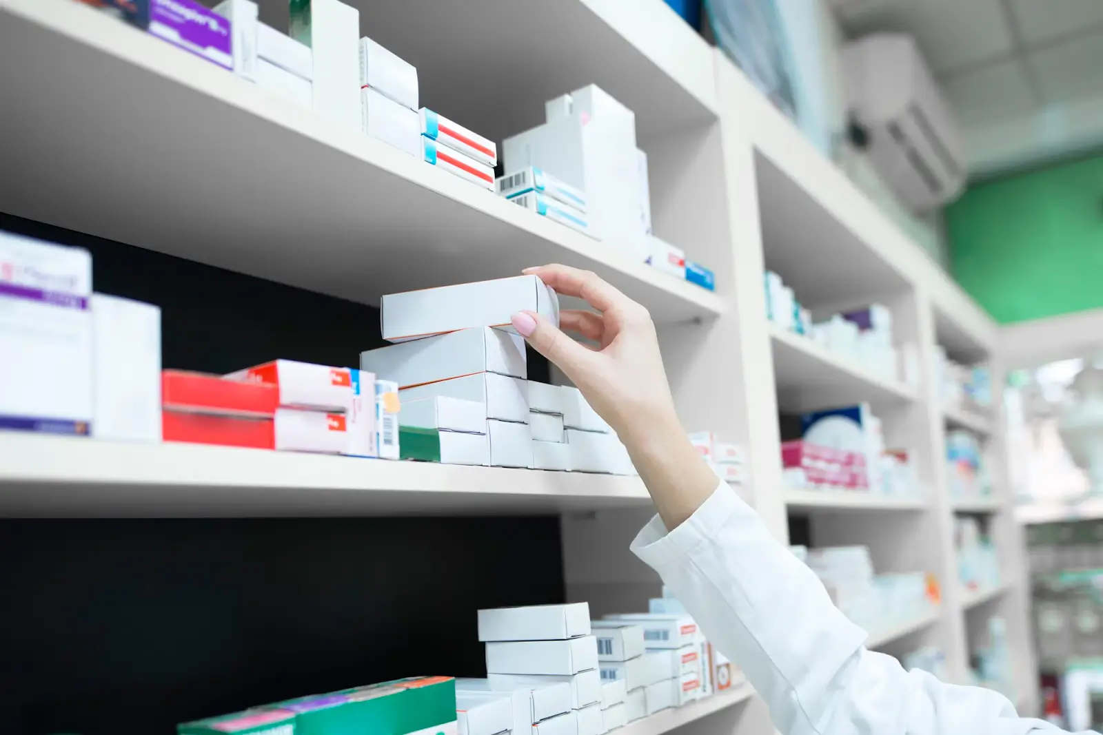 A person in a white coat reaches for a box on a pharmacy shelf filled with various medications in boxes. The background shows a well-organized shelving system with neatly arranged pharmaceutical products.