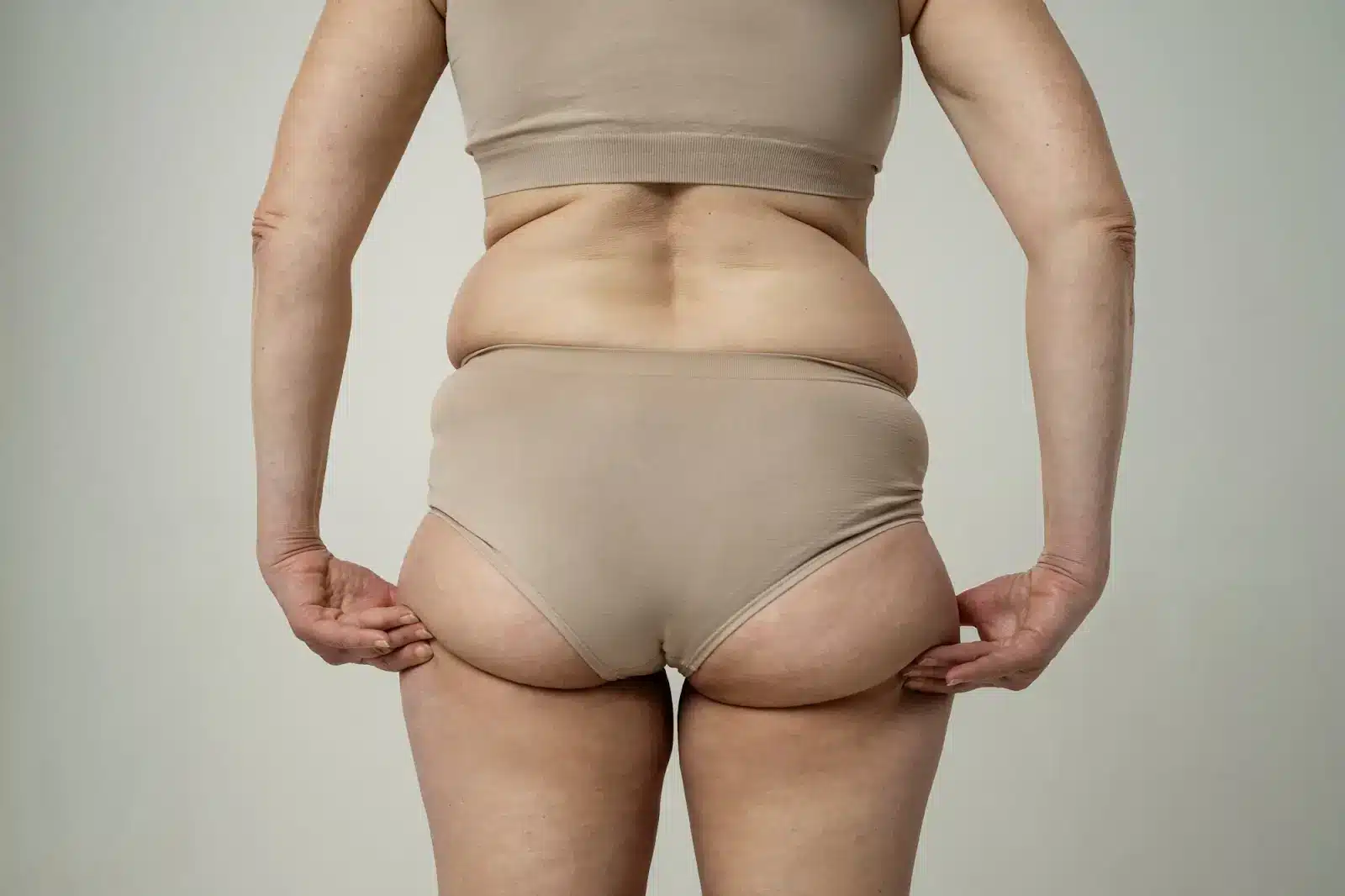 A woman wearing beige underwear and a matching top is shown from behind, standing against a plain background and gently holding the sides of her underwear. Her back and waist are visible.