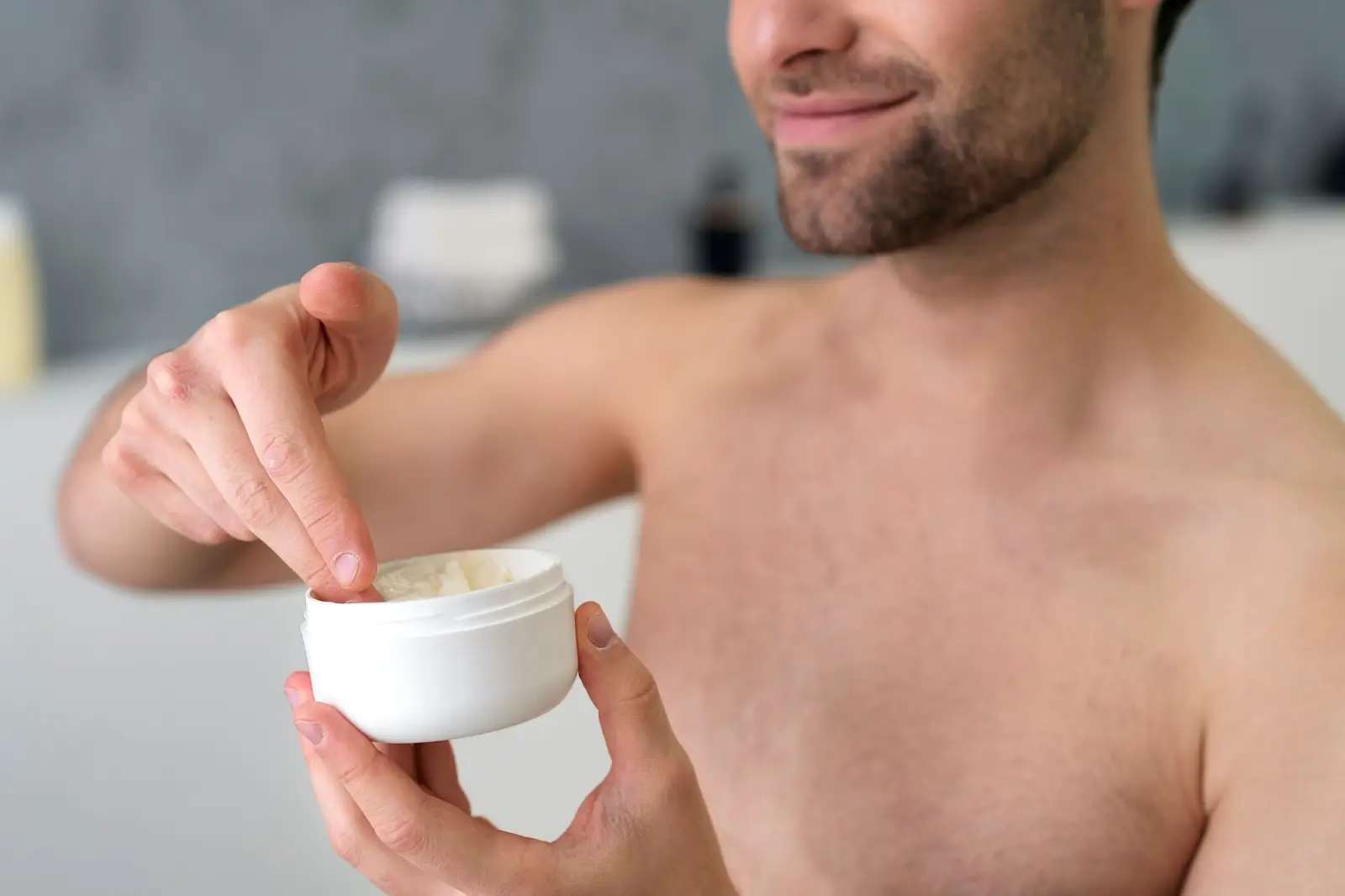A shirtless person with short hair smiling, applying a cream from a white jar onto their fingers. The background is blurred, suggesting a bathroom setting.