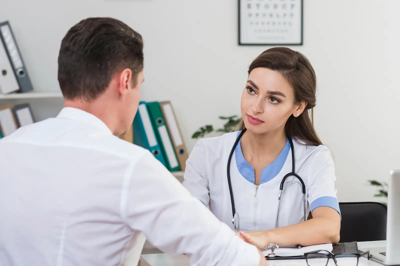 A female doctor with a stethoscope listens attentively to a male patient in an office setting. They are seated at a desk near shelves with binders and framed artwork, creating a professional atmosphere.
