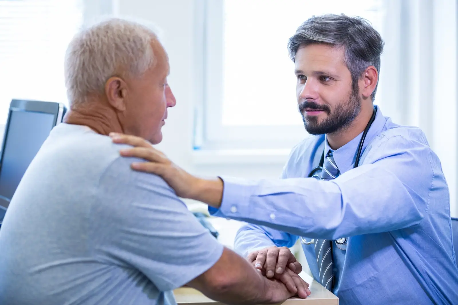 A doctor with a stethoscope around his neck gently touches the shoulder of an older man in a gray shirt. They are sitting at a desk in a bright medical office, having a conversation.