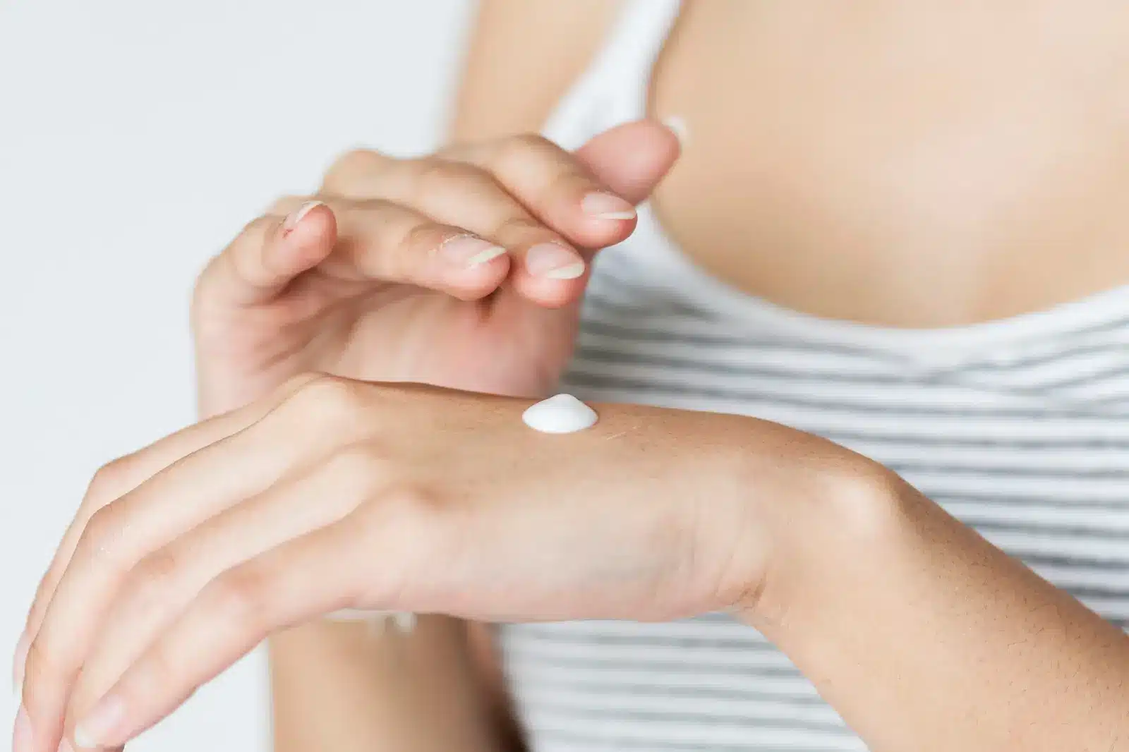 A person applies a dollop of white cream to the back of their hand. They are wearing a white and gray striped tank top. The background is plain white.