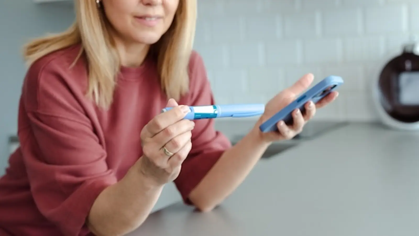 A woman holds a blue insulin pen in one hand and a smartphone in the other, sitting at a kitchen counter, with only her face partially visible.