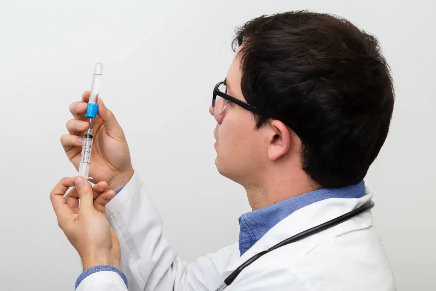 A person wearing a white lab coat and stethoscope prepares a syringe by drawing liquid from a vial, against a plain white background.
