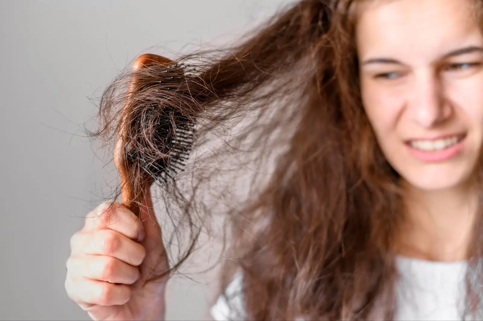 A woman with long, tangled hair struggles to brush it, showing frustration as her hair is caught in the bristles of a wooden hairbrush.