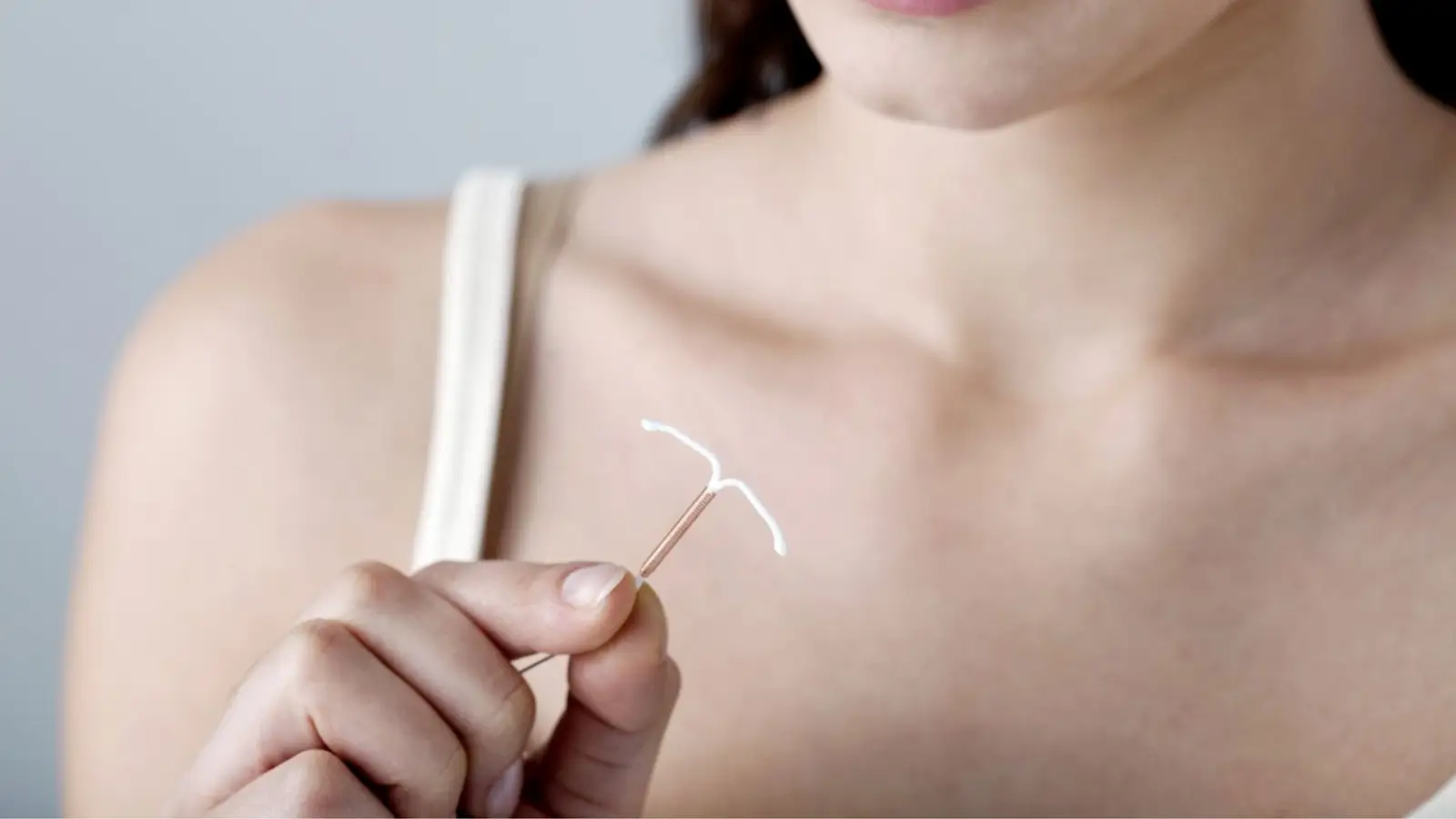 A woman in a tank top holds a copper intrauterine device (IUD) between her fingers, with her face partially visible and the focus on the IUD.