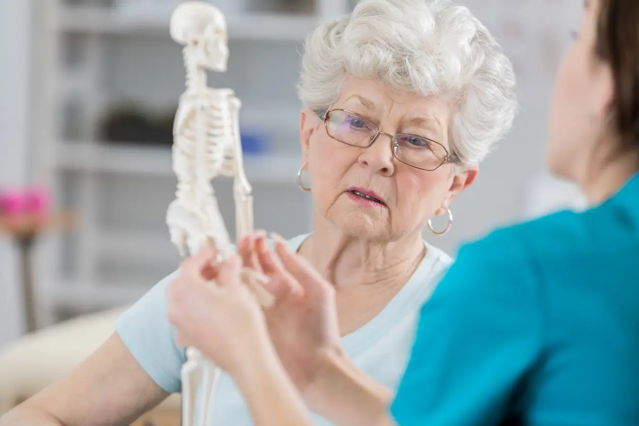 An older woman with gray hair and glasses listens attentively to a healthcare professional explaining something using a small human skeleton model.