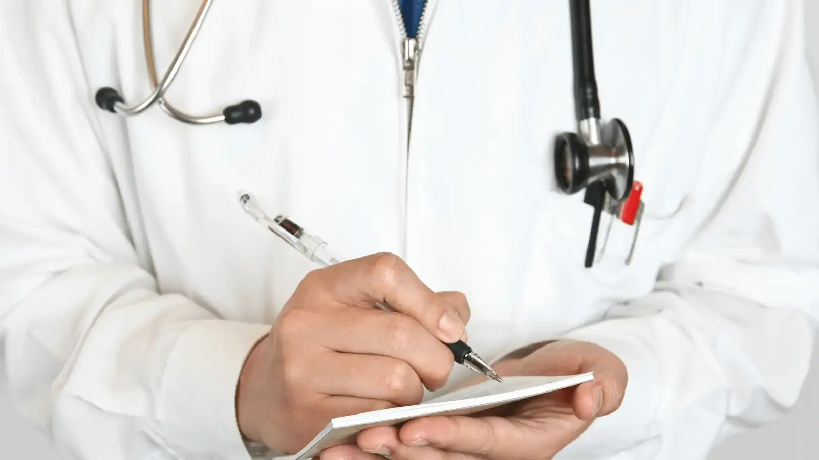 A doctor wearing a white coat and stethoscope writes on a notepad while holding it in one hand, focusing on the task.