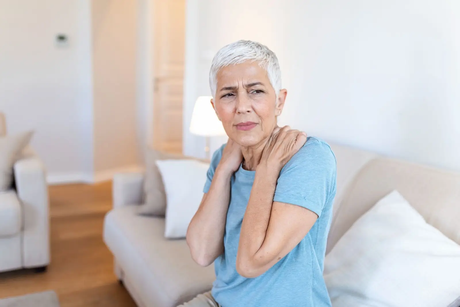 An older woman with short gray hair sits on a couch, holding her shoulders and appearing to be in discomfort or pain, possibly from muscle tension or stiffness, in a brightly lit living room.