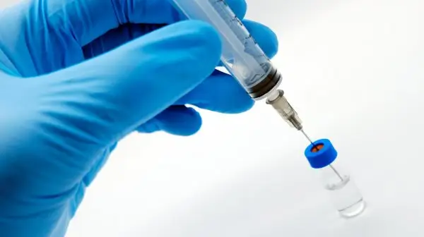 A gloved hand holds a syringe, drawing liquid from a small vial with a blue cap, against a white background.