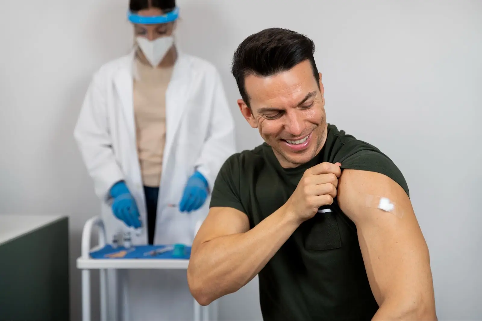 A smiling man in a dark green shirt shows his upper arm with a small bandage after getting vaccinated. In the background, a healthcare worker in protective gear stands by a medical cart with supplies.