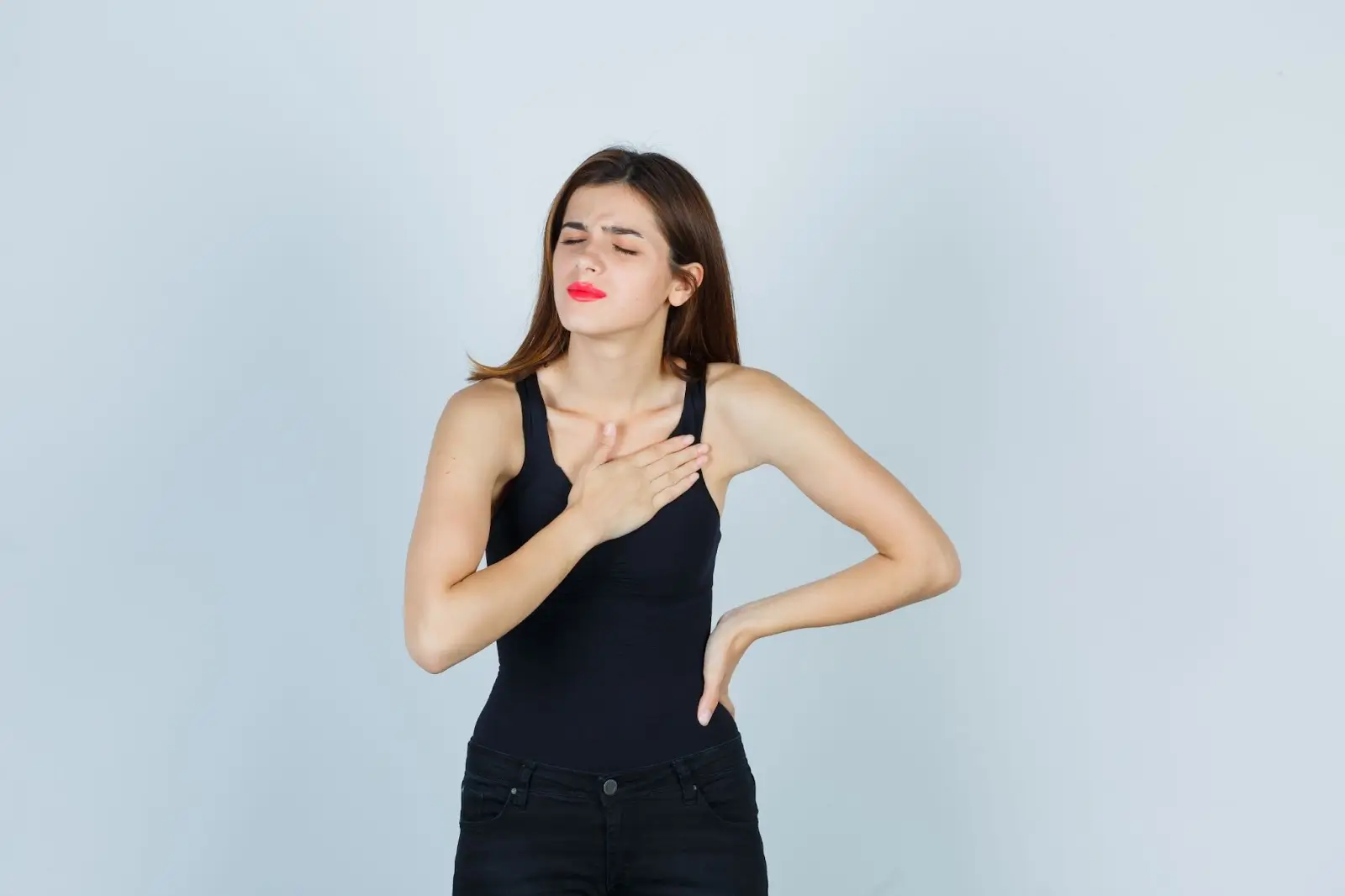 A young woman in a black tank top holds her chest with one hand and her side with the other, eyes closed and face showing discomfort, suggesting she is experiencing chest pain.