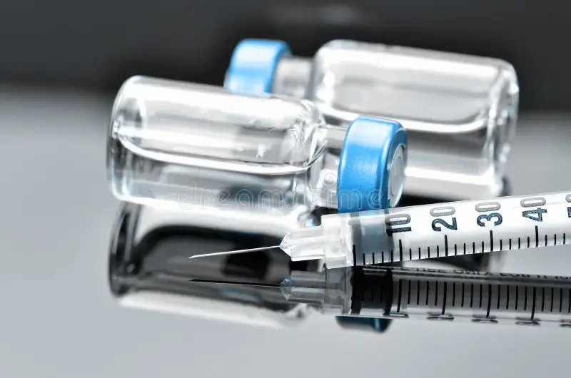 Close-up of a syringe with a needle next to two clear glass vials sealed with blue caps, all reflected on a shiny surface.