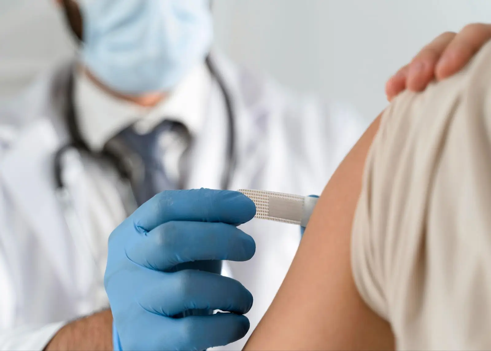 A healthcare professional wearing a mask and blue gloves administers a vaccine injection into a person's upper arm. The person is dressed in a short-sleeved shirt and is gently holding their shoulder.